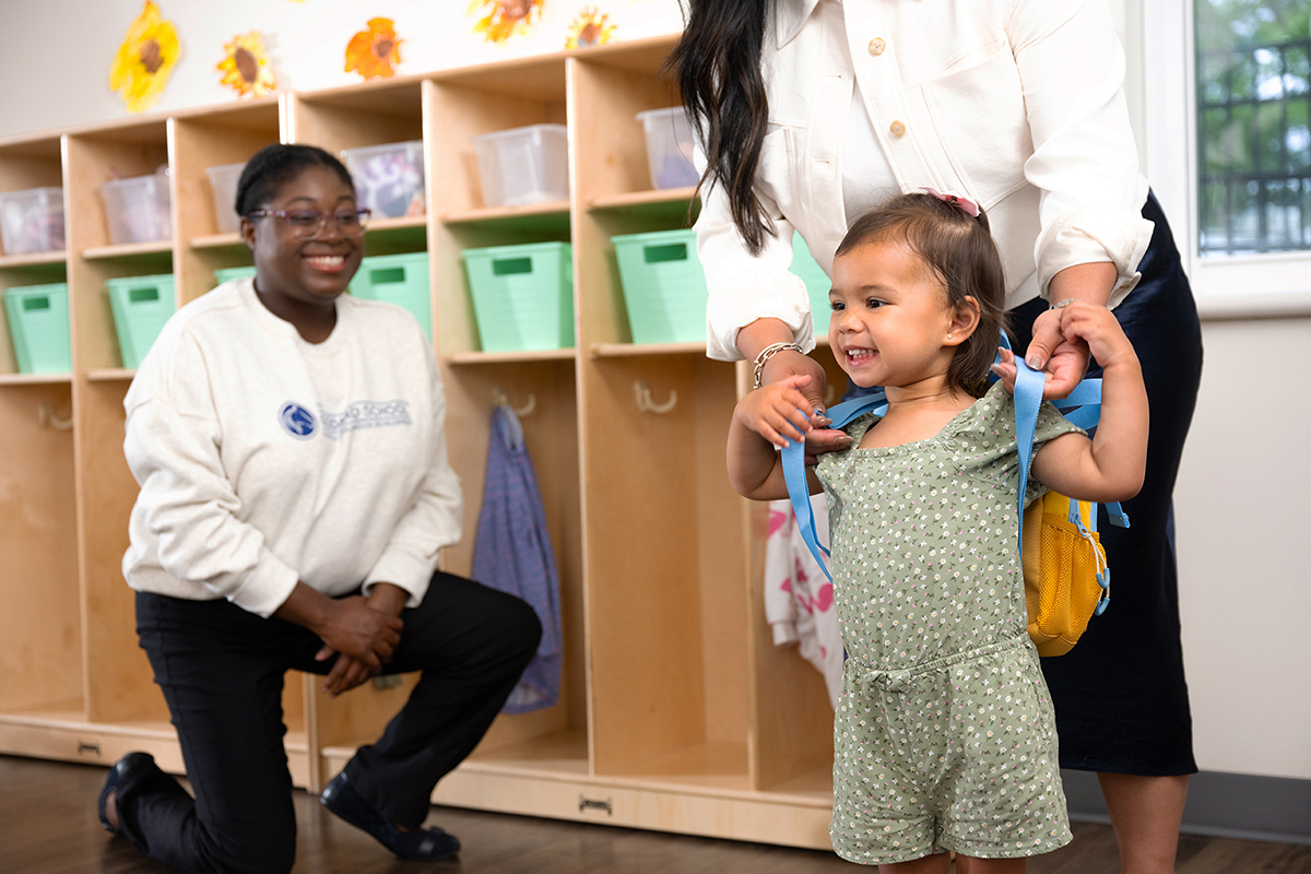 A mom removing her daughter's backpack in a preschool classroom as a smiling Goddard School teacher looks on
