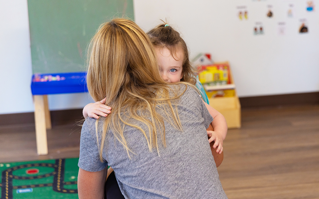 A shy little girl hugging her mother in a preschool classroom.