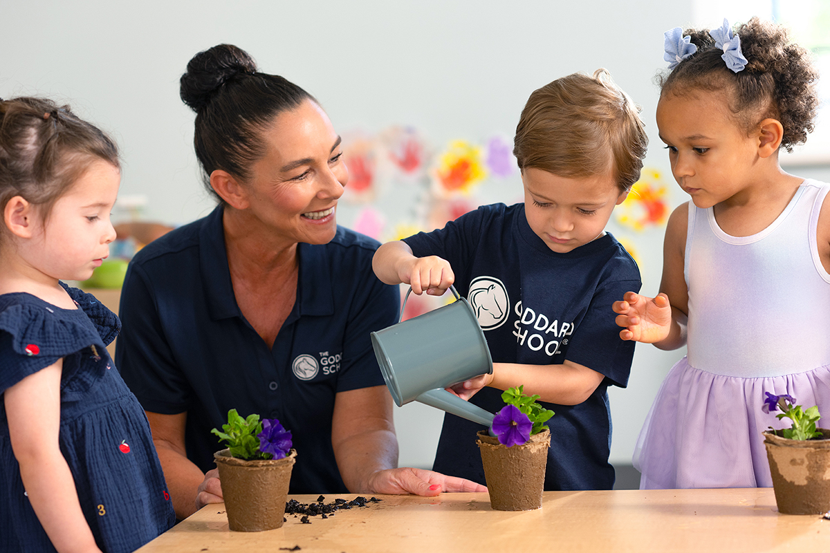 A teacher and three students watering plants at a table in a preschool classroom