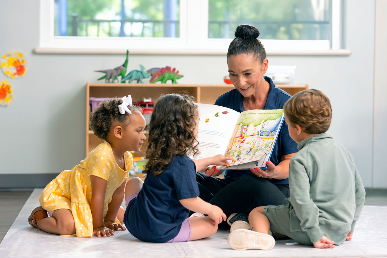 A teacher reading Curious Blueberry the Carousel Horse to three children in a Goddard School classroom.