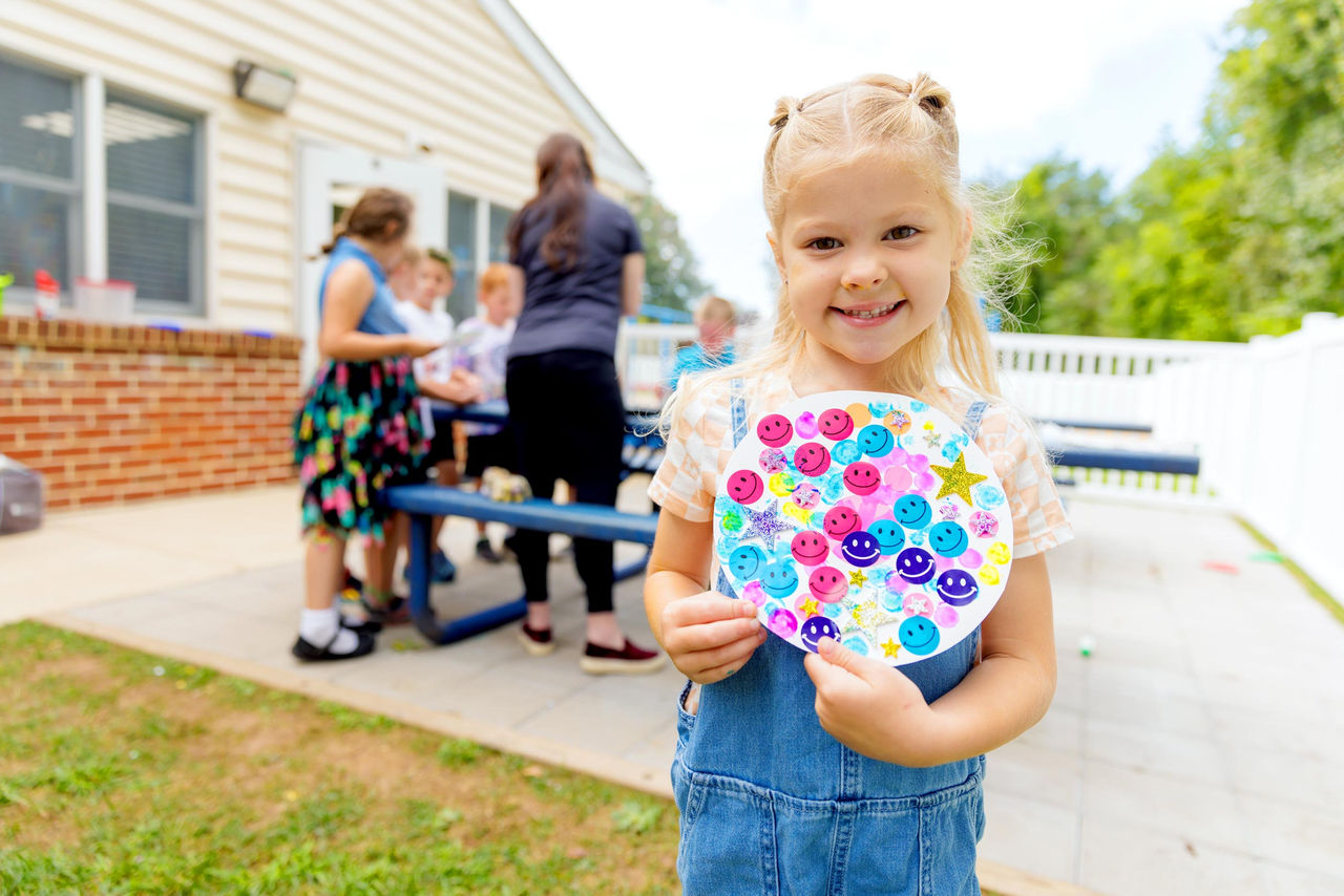 A smiling child proudly showing off her art project on an outdoor patio