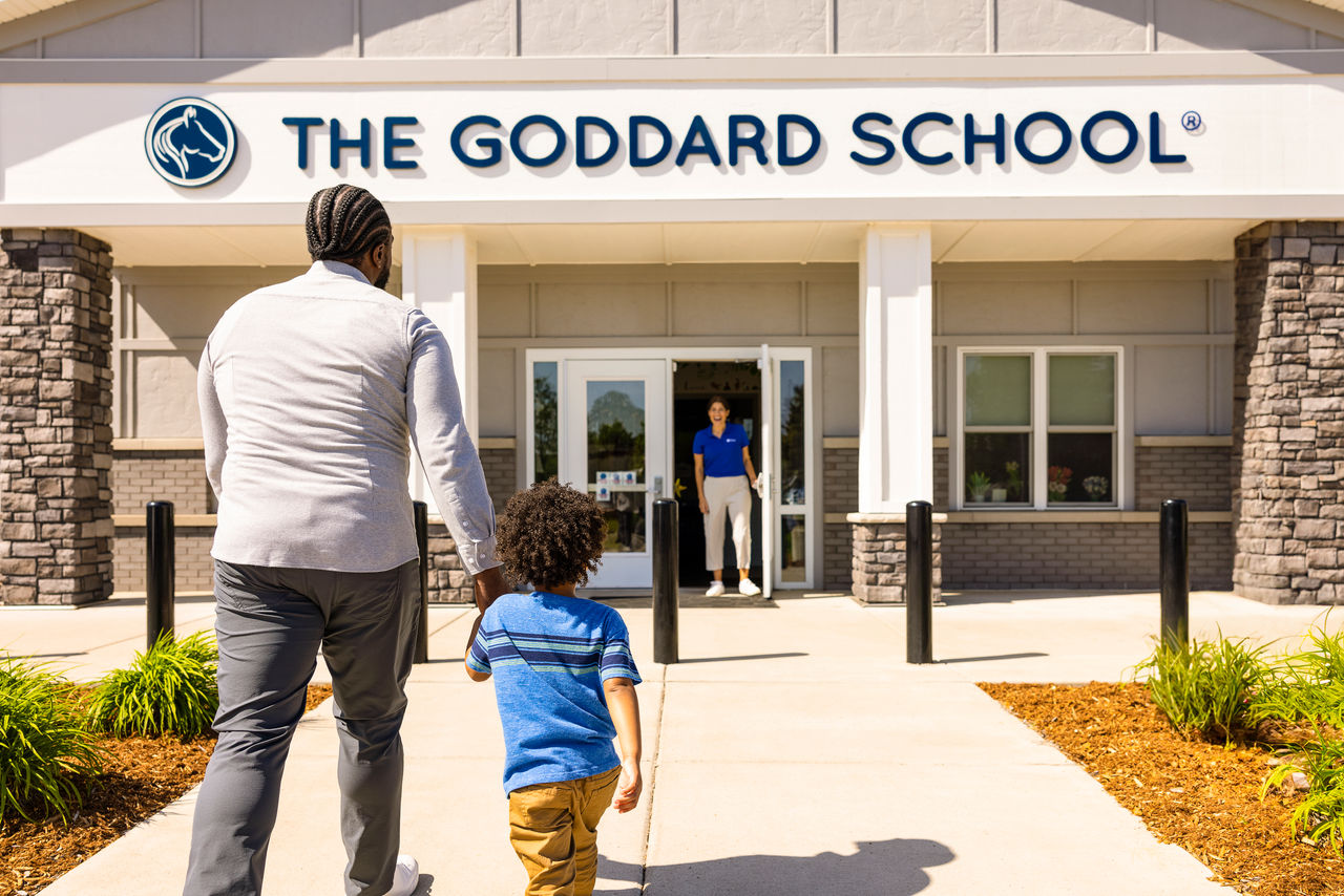 A father and son holding hands walking up to a Goddard School entryway where a teacher is waiting to greet them.